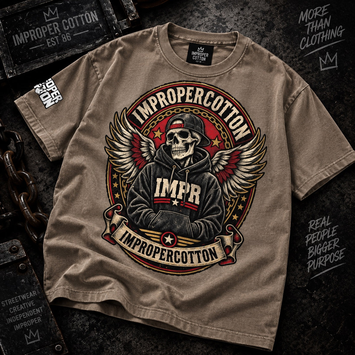 A tan graphic t-shirt featuring a central circular design of a winged skeleton wearing a cap and hoodie with 'IMPR' on the chest, surrounded by the words 'IMPROPERCOTTON'. The t-shirt is styled in a flat-lay on a dark textured background with vintage crates and chains.