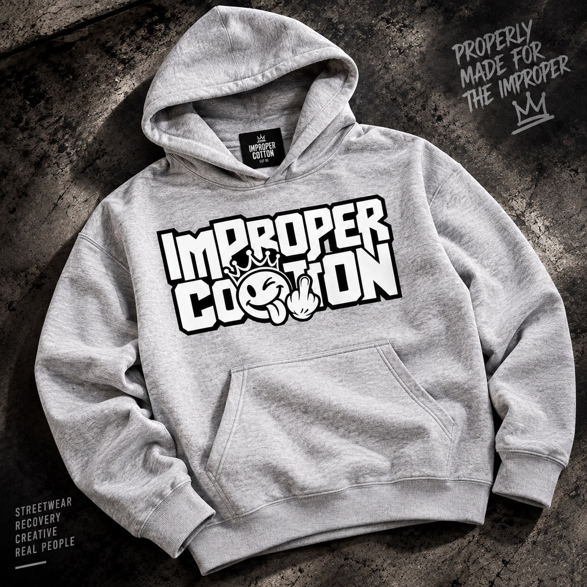 A heather gray pullover hoodie laid flat on a dark textured surface, featuring a large white graphic print on the chest that reads “IMPROPER COPTION” with a small illustrated face and crown. A black neck label is visible, and decorative text appears in the corners.