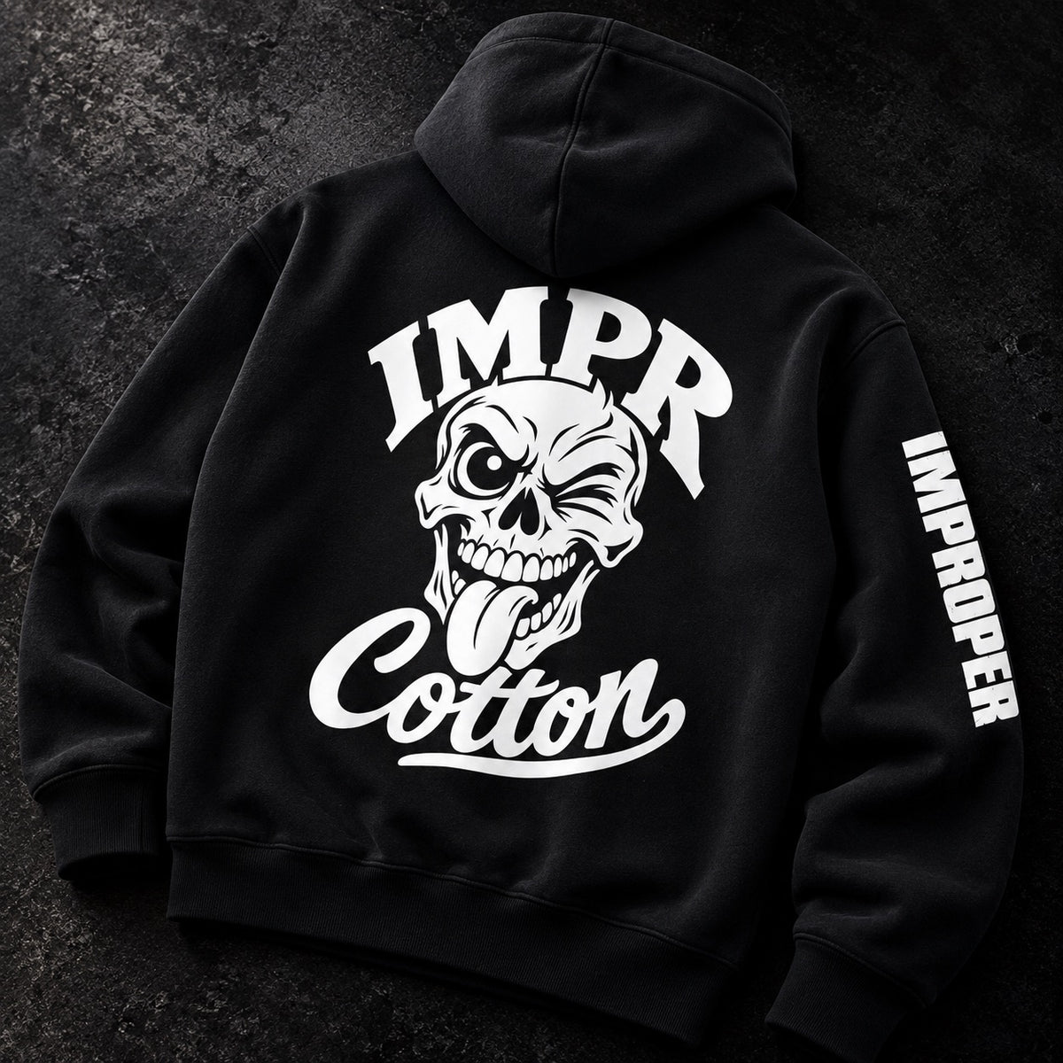Black pullover hoodie laid flat on a dark textured surface, showing the back with a large white graphic of a skull and the words “IMPR Cotton,” plus vertical “IMPROPER” text on the sleeve.