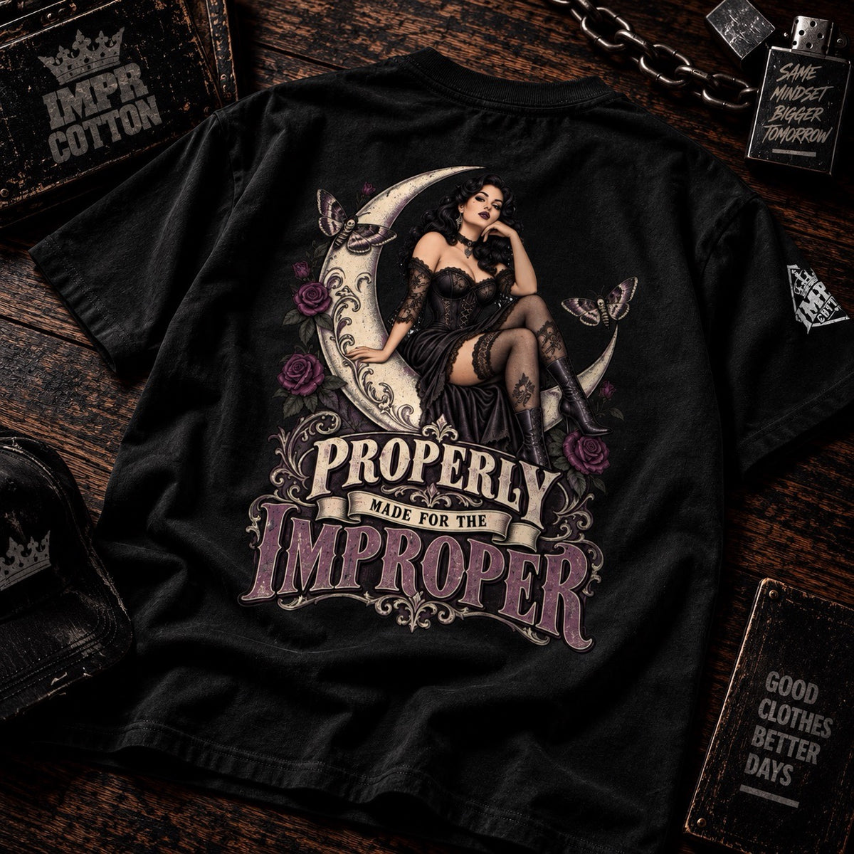 A black graphic t-shirt laid flat on a dark wooden background, featuring a vintage-style illustration of a woman in a black dress sitting on a crescent moon with roses, and the text 'PROPERLY MADE FOR THE IMPROPER'.