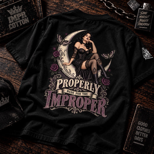 A black graphic t-shirt laid flat on a dark wooden background, featuring a vintage-style illustration of a woman in a black dress sitting on a crescent moon with roses, and the text 'PROPERLY MADE FOR THE IMPROPER'.