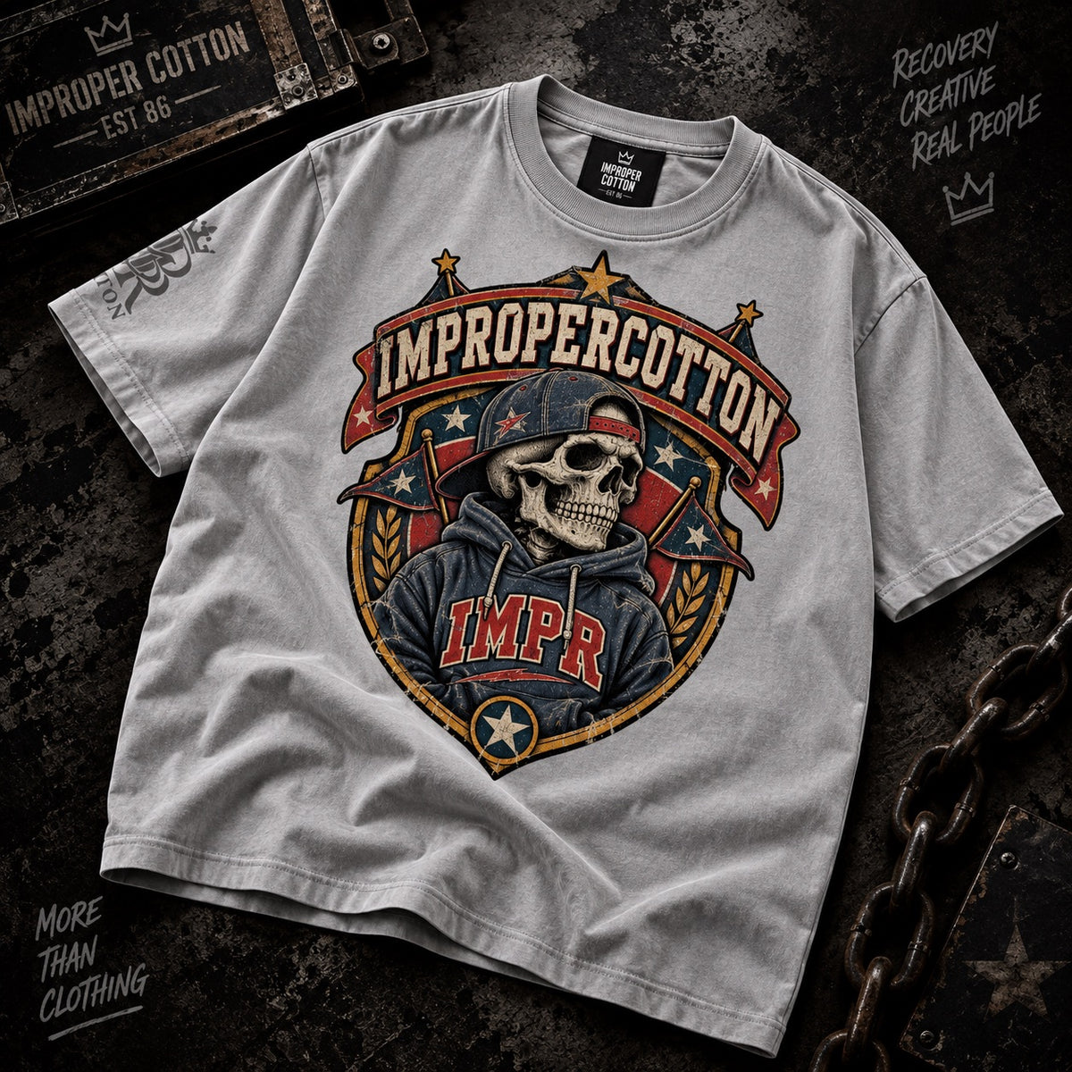 A light gray heather t-shirt with a large graphic print of a skeleton wearing a cap and hoodie, with the text 'IMPROPERCOTTON' and 'IMPR'. The shirt is laid flat on a dark textured background with rustic props.