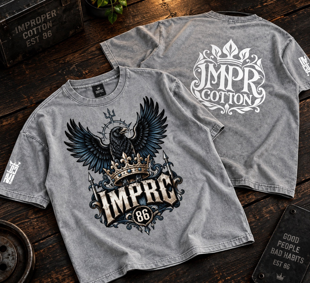 Two heather gray graphic T-shirts displayed on a dark wooden surface. The front shirt features a large black and white eagle with a gold crown and the text 'IMPRC 86'.