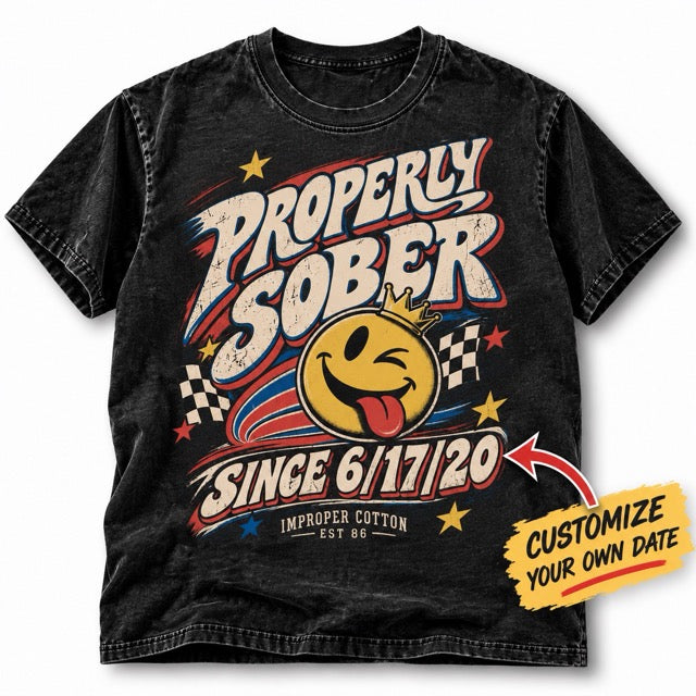 A black acid-wash graphic t-shirt featuring a retro racing design with the text 'PROPERLY SOBER SINCE 6/17/20' and a winking yellow smiley face wearing a crown. A yellow 'CUSTOMIZE YOUR OWN DATE' arrow sticker is overlaid on the bottom right.