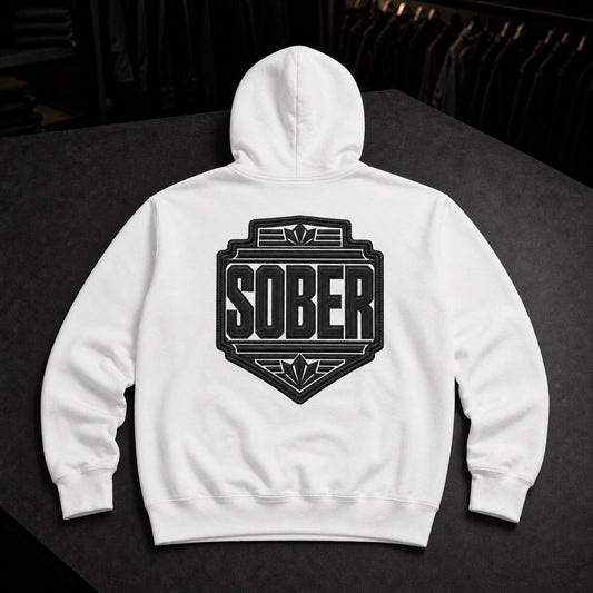 A white pullover hoodie shown from the back, featuring a large black shield-style graphic with the word 'SOBER' in bold letters. The hoodie is laid flat on a dark surface with a blurred clothing rack in the background.
