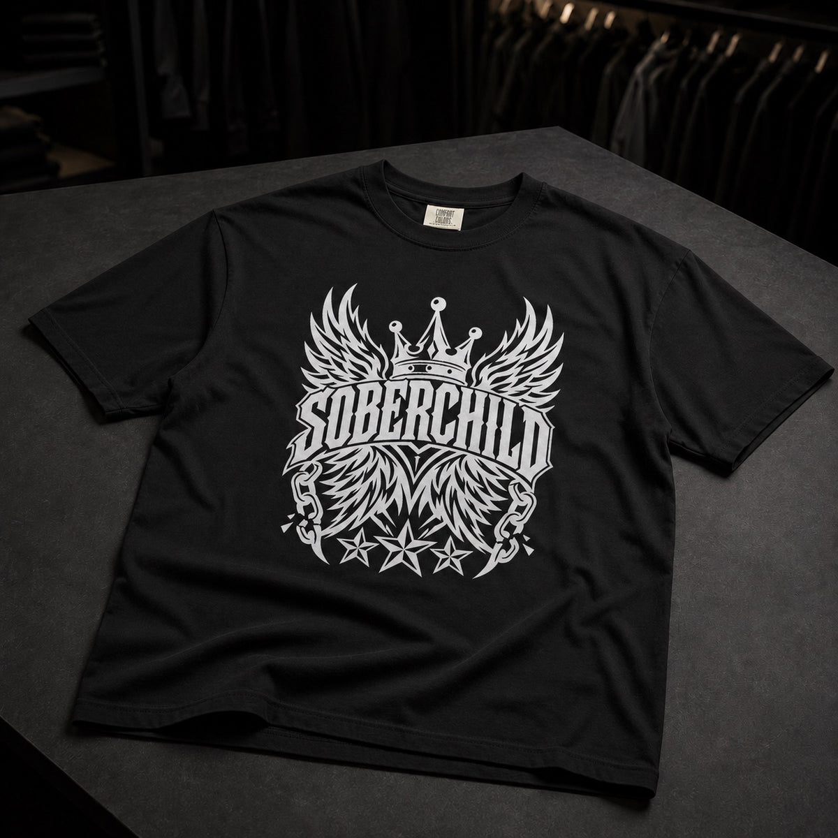 A black short-sleeve t-shirt with a large white graphic on the front featuring wings, a crown, and the text 'SOBERCHILD' and 'MM', displayed on a dark gray surface.