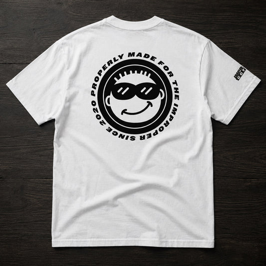 A white short-sleeve t-shirt laid flat on a dark wood background, featuring a large black circular graphic on the back with a smiley face wearing sunglasses and the text 'PROPERLY MADE FOR THE IMPROPER SINCE 2020'. A small black logo is printed on the left sleeve.