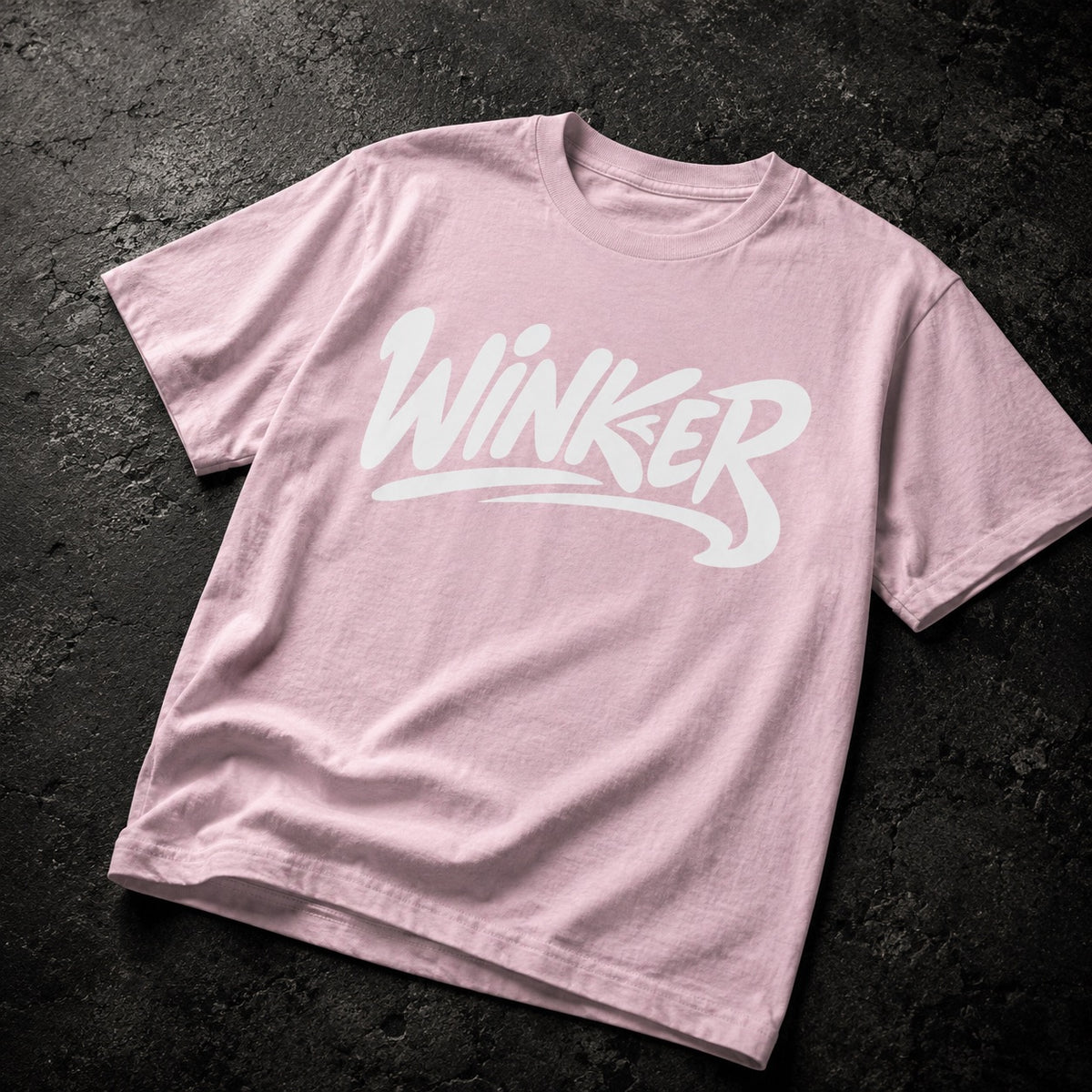 A light pink crewneck t-shirt with a white stylized 'WINKER' graphic printed across the chest, laid flat on a dark textured background.