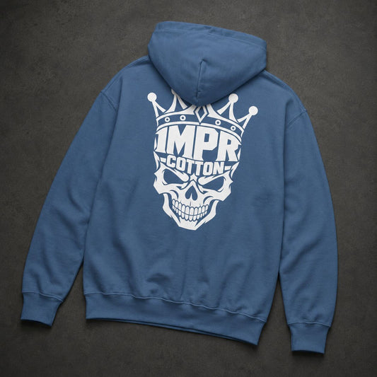 A blue pullover hoodie shown from the back, featuring a large white graphic of a skull wearing a crown with the text 'IMPR COTTON' printed across it, laid flat on a dark textured background.