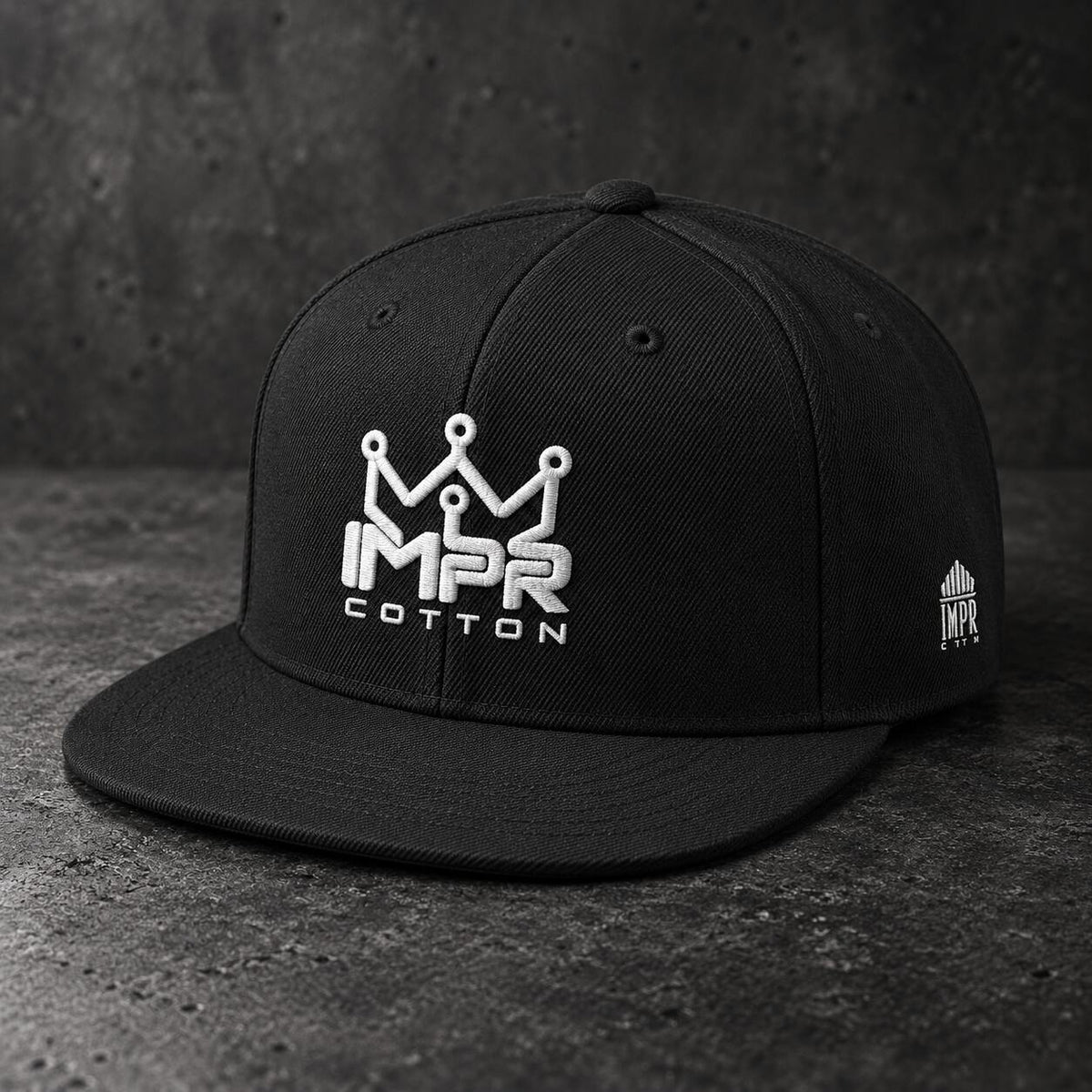 A black flat-brim snapback cap with a white embroidered crown logo and the text 'IMPR COTTON' on the front, shown on a dark textured surface.