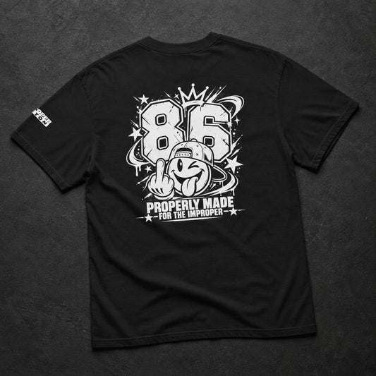 A black short-sleeve t-shirt laid flat on a dark textured background, showing a large white graphic print on the back with the number 86, a cartoon character, stars, and the text 'PROPERLY MADE FOR THE IMPROPER'. A small white logo is visible on the left sleeve.
