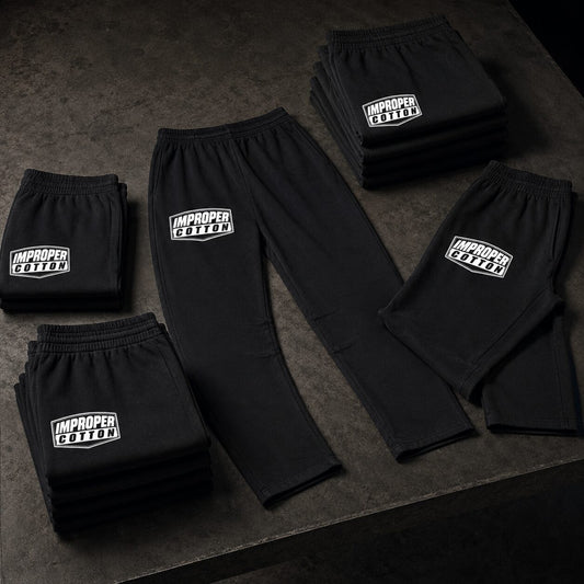 Black sweatpants laid out on a dark textured surface, with several folded pairs arranged around one unfolded pair. Each pair has a white rectangular 'IMPROPER COLLECTIVE' logo patch on the upper thigh.