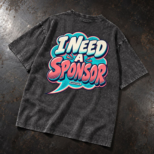 A dark gray acid-wash t-shirt laid flat on a textured dark gray surface, featuring a large graphic on the back with the text 'I NEED A SPONSOR' in a colorful comic-style speech bubble.