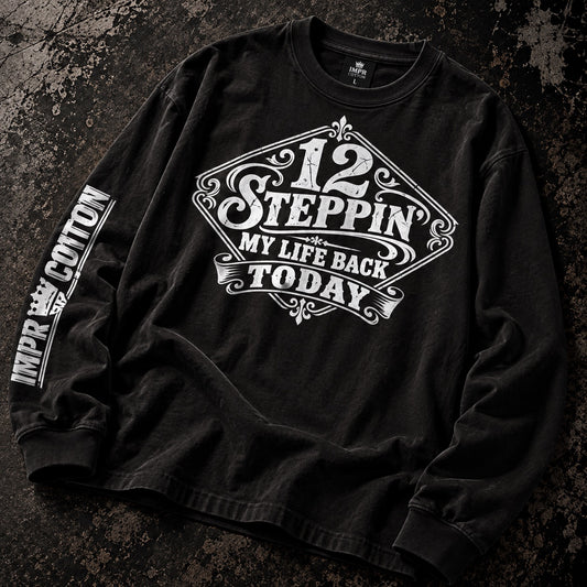 A black long-sleeve t-shirt with a distressed wash, featuring a white graphic print on the front that reads '12 STEPPIN' MY LIFE BACK TODAY' and 'IMPR COTTON' printed vertically on the sleeve, laid flat on a dark textured background.