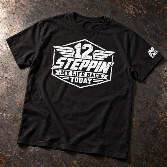 A black short-sleeve graphic t-shirt laid flat on a textured, dark brown surface. The shirt features a large white distressed graphic on the front reading '12 STEPPIN MY LIFE BACK TODAY' with wing-like details, and a smaller white graphic on the right sleeve.