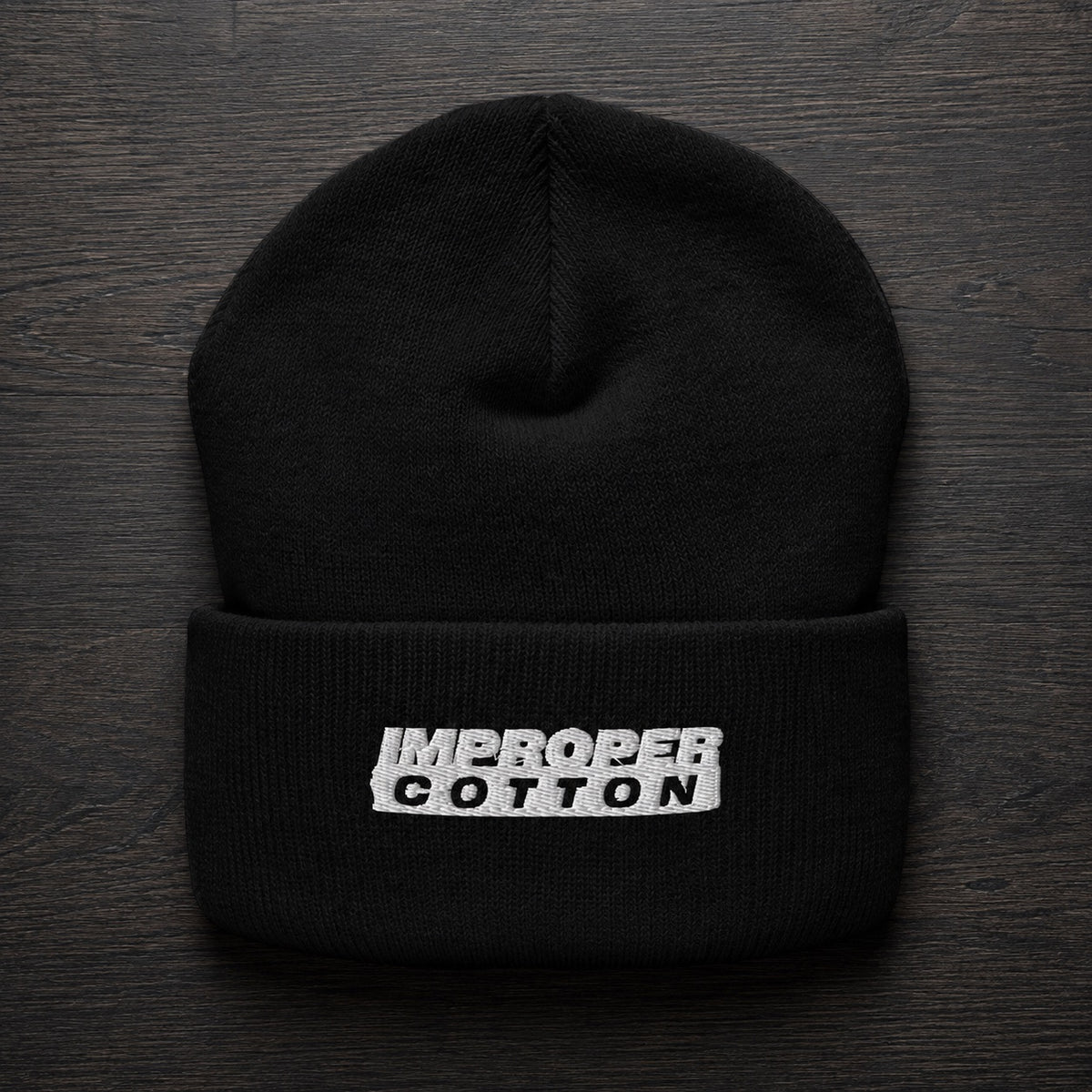 A black knit beanie hat with a folded cuff, featuring a white embroidered rectangular patch with the words 'IMPROPER COTTON' in black. The hat is photographed flat on a dark wood background.