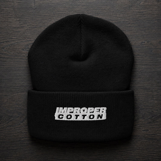 A black knit beanie hat with a folded cuff, featuring a white embroidered rectangular patch with the words 'IMPROPER COTTON' in black. The hat is photographed flat on a dark wood background.