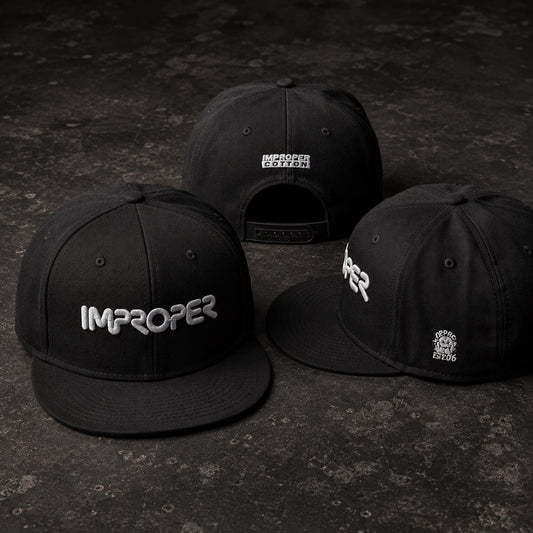 Three black flat-brim snapback caps arranged on a dark textured surface. The front cap shows the word 'IMPROPER' in white across the front, the right cap shows part of the same white logo on the side, and the back cap shows the back of the brim with additional small white text.