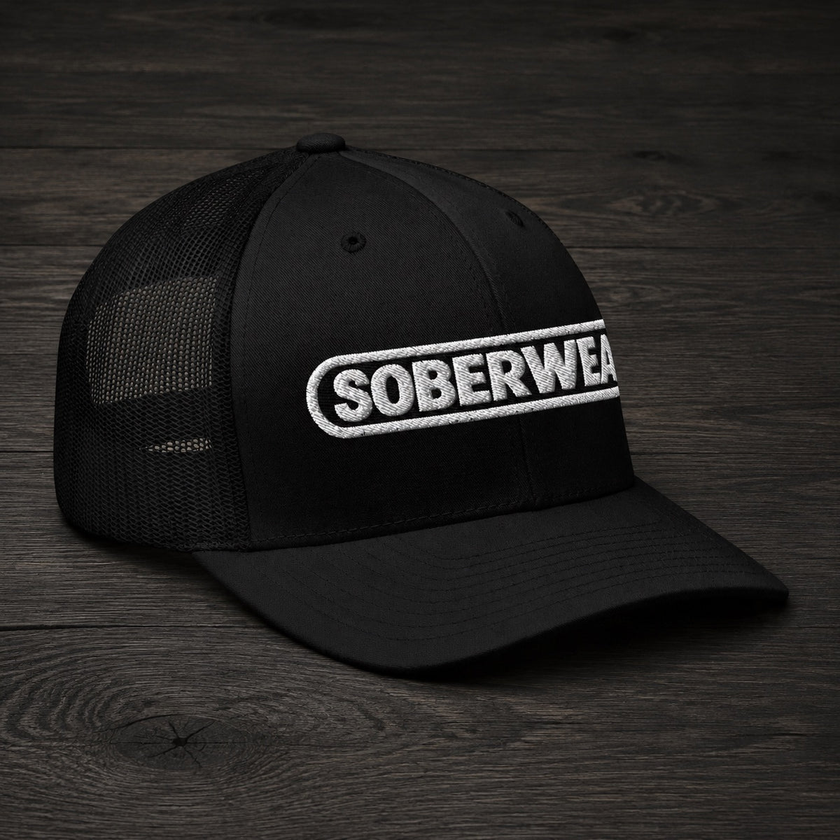 A black trucker-style cap with a curved brim and black mesh back panels, featuring a white embroidered 'SOBERWEAR' logo on the front, displayed on a dark wood surface.