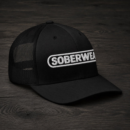 A black trucker-style cap with a curved brim and black mesh back panels, featuring a white embroidered 'SOBERWEAR' logo on the front, displayed on a dark wood surface.