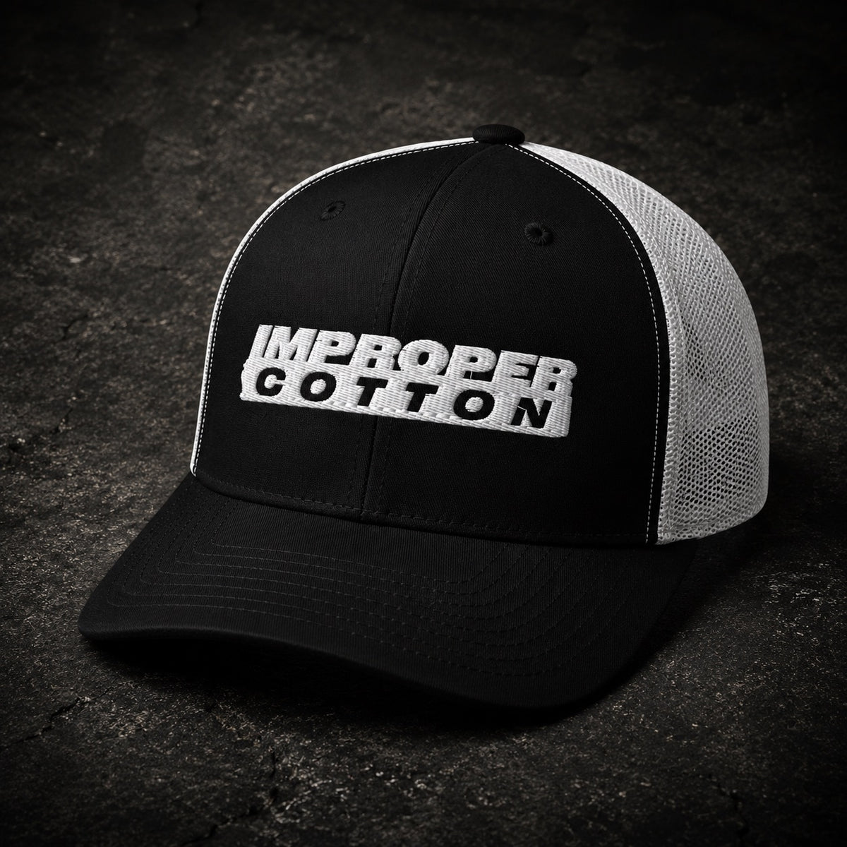 A black and white trucker hat with 'IMPROPER COTTON' embroidered on the front, set against a dark textured background.