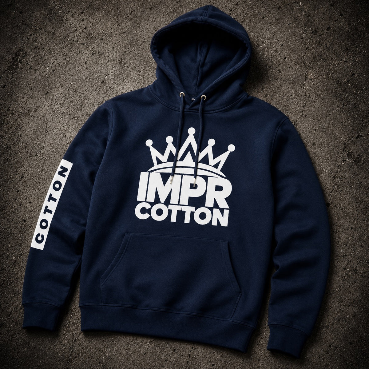 A navy blue pullover hoodie laid flat on a textured dark gray surface. The hoodie features a large white graphic on the chest with a crown above the words 'IMPR COTTON', and a vertical white 'COTTON' label on the right sleeve.