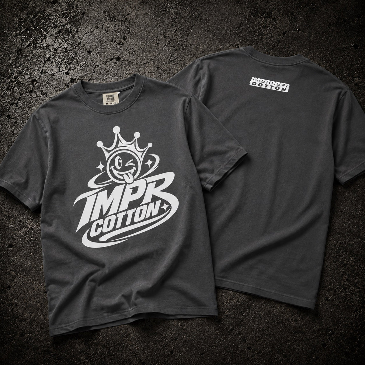 Two dark charcoal gray t-shirts displayed flat on a textured dark background, showing the front and back designs. The front features a large white graphic of a cartoon face inside a crown with the text 'IMPR COTTON+'.