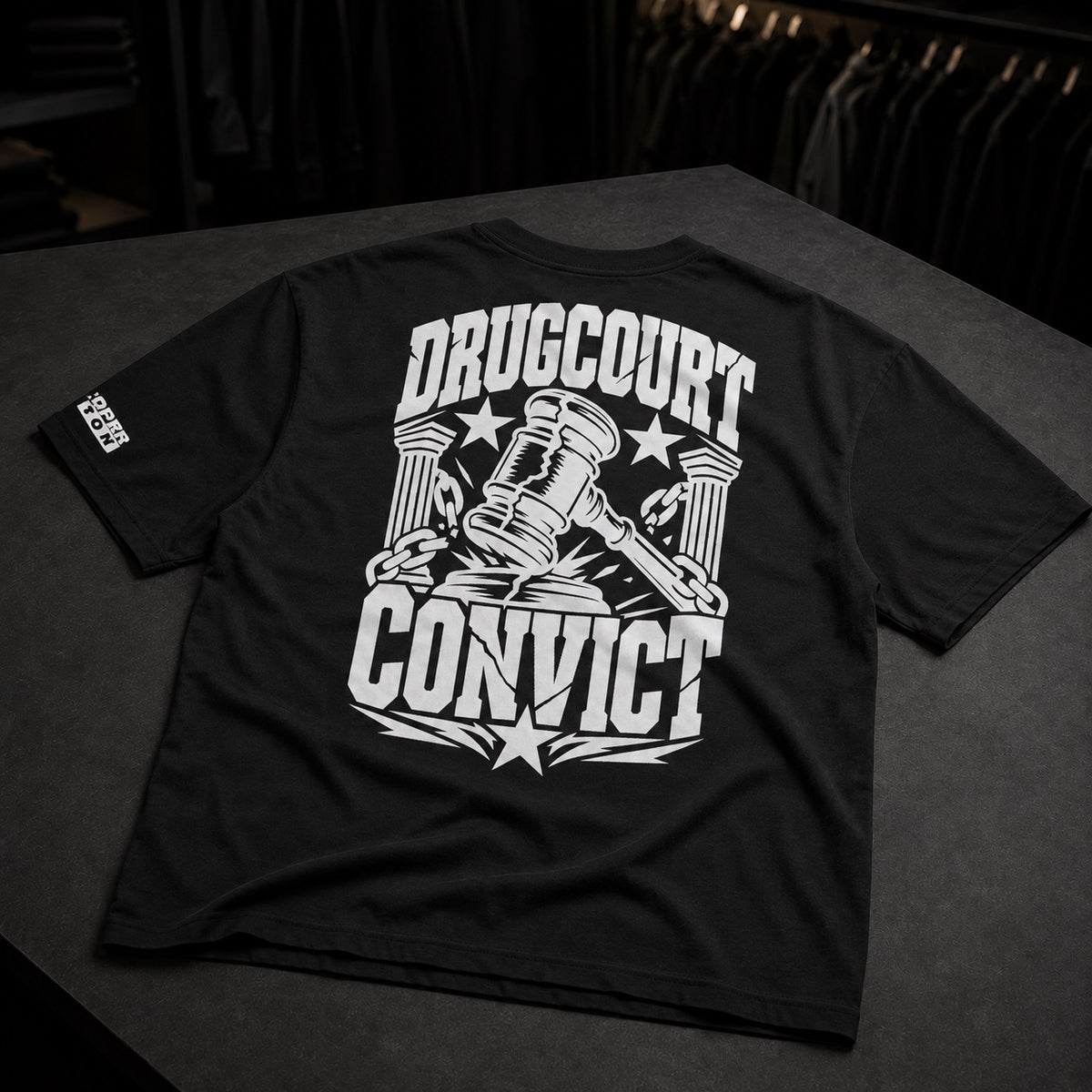 A black short-sleeve t-shirt laid flat on a dark surface, showing the back with a large white graphic that reads 'DRUGCOURT CONVICT' around a gavel and columns design. A small white logo is visible on the left sleeve.