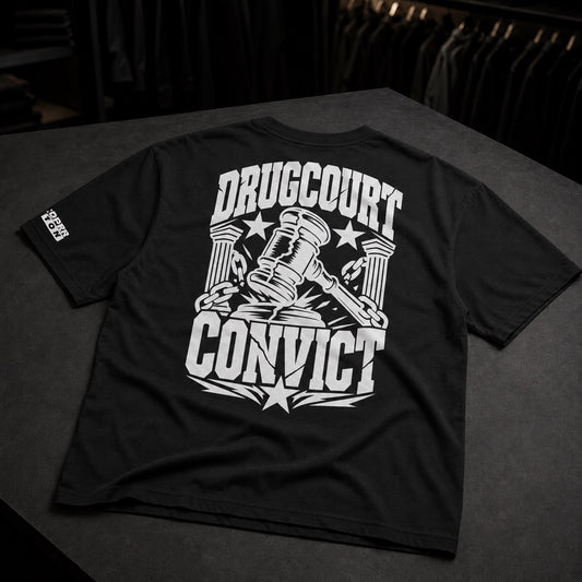 A black short-sleeve t-shirt laid flat on a dark surface, showing the back with a large white graphic that reads 'DRUGCOURT CONVICT' around a gavel and columns design. A small white logo is visible on the left sleeve.