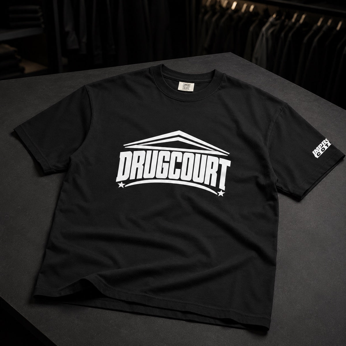 A black short-sleeve t-shirt with a bold white 'DRUGCOURT' graphic printed across the chest, displayed on a dark gray table with a rack of clothes in the background.