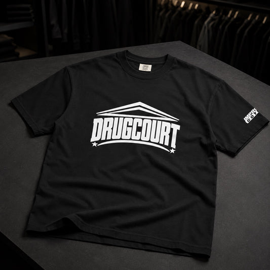 A black short-sleeve t-shirt with a bold white 'DRUGCOURT' graphic printed across the chest, displayed on a dark gray table with a rack of clothes in the background.