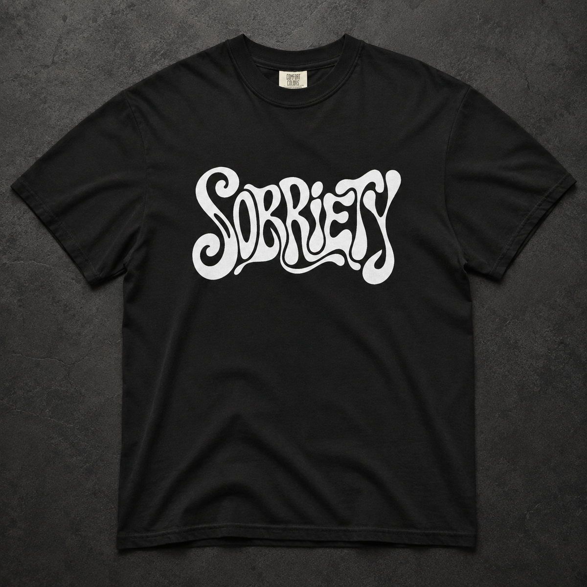 A black short-sleeve t-shirt with the word 'SOBRIETY' printed across the chest in a stylized white bubble-letter font, laid flat on a dark textured background.