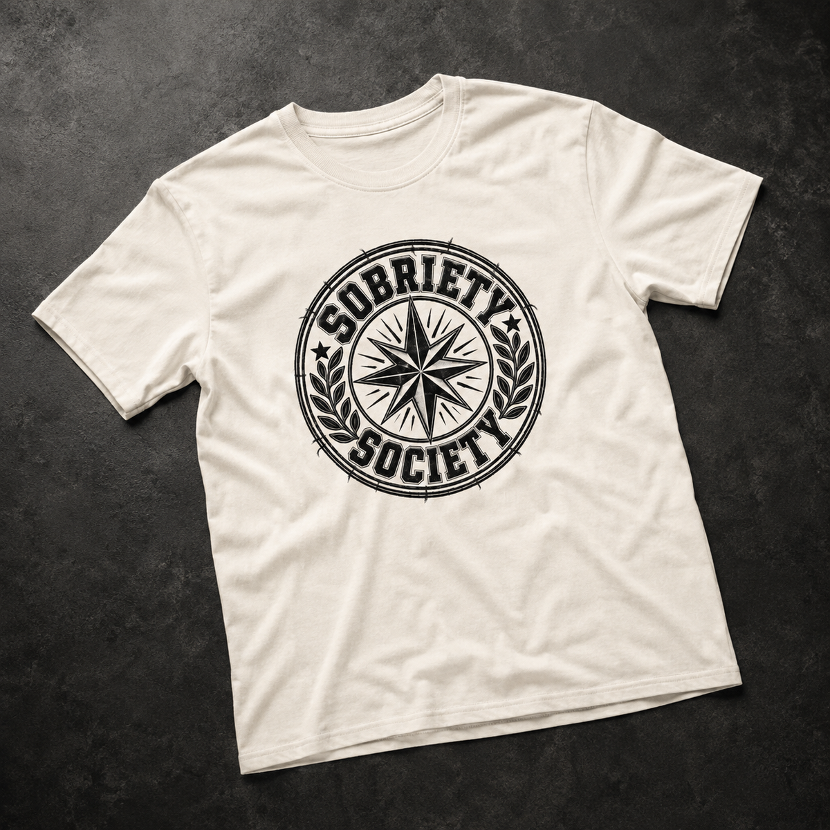 An off-white crewneck t-shirt with a black distressed circular 'Sobriety Society' graphic on the chest, laid flat on a dark textured background.