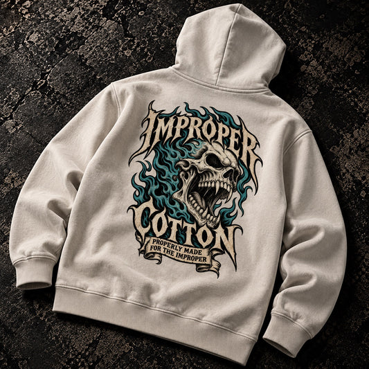 An off-white hooded sweatshirt laid flat on a dark textured surface, showing the back graphic. The design features a large skull surrounded by blue and black flames, with the words 'IMPROPER COTTON' in a stylized font and a smaller banner reading 'IMPERially MADE FOR THE IMPROPER'.