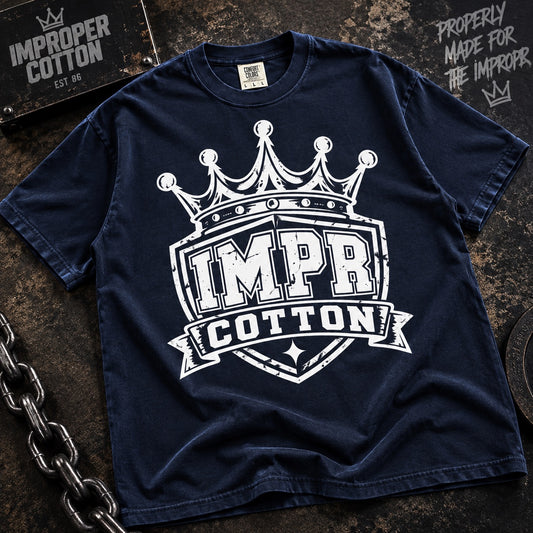 A navy blue short-sleeve t-shirt with a large white distressed graphic of a crown and the words 'IMPR COTTON' on the front, laid flat on a dark textured surface with chain and sign props.
