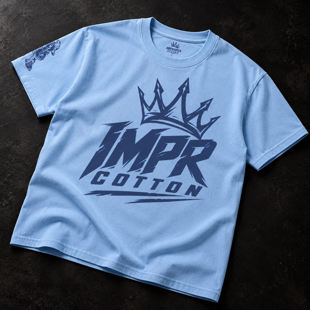 A light blue crewneck t-shirt with a dark navy graphic on the front featuring a crown and the text 'IMPR COTTON'. The t-shirt is laid flat on a dark textured background.