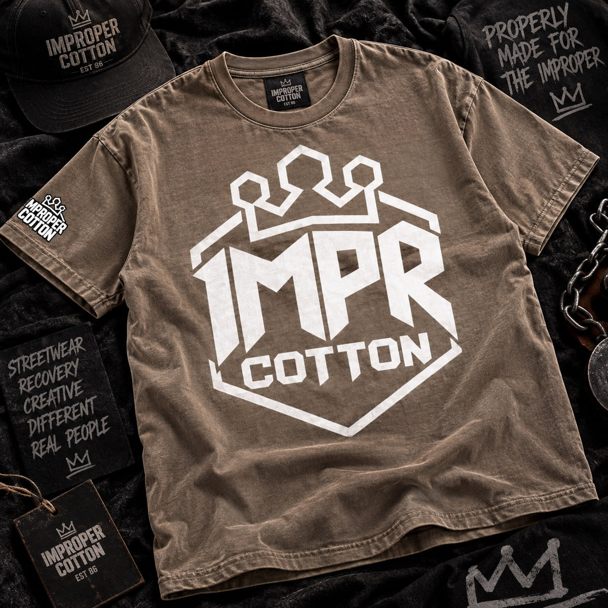 A brown short-sleeve graphic T-shirt laid flat on a dark textured surface, featuring a large white 'IMPR COTTON' shield logo with a crown above the text. Matching branded black caps, patches, and tags are arranged around it in a styled flat-lay.