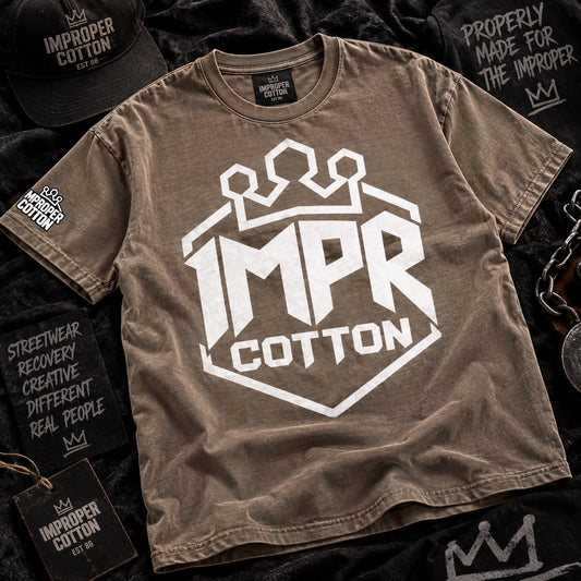A brown short-sleeve graphic T-shirt laid flat on a dark textured surface, featuring a large white 'IMPR COTTON' shield logo with a crown above the text. Matching branded black caps, patches, and tags are arranged around it in a styled flat-lay.