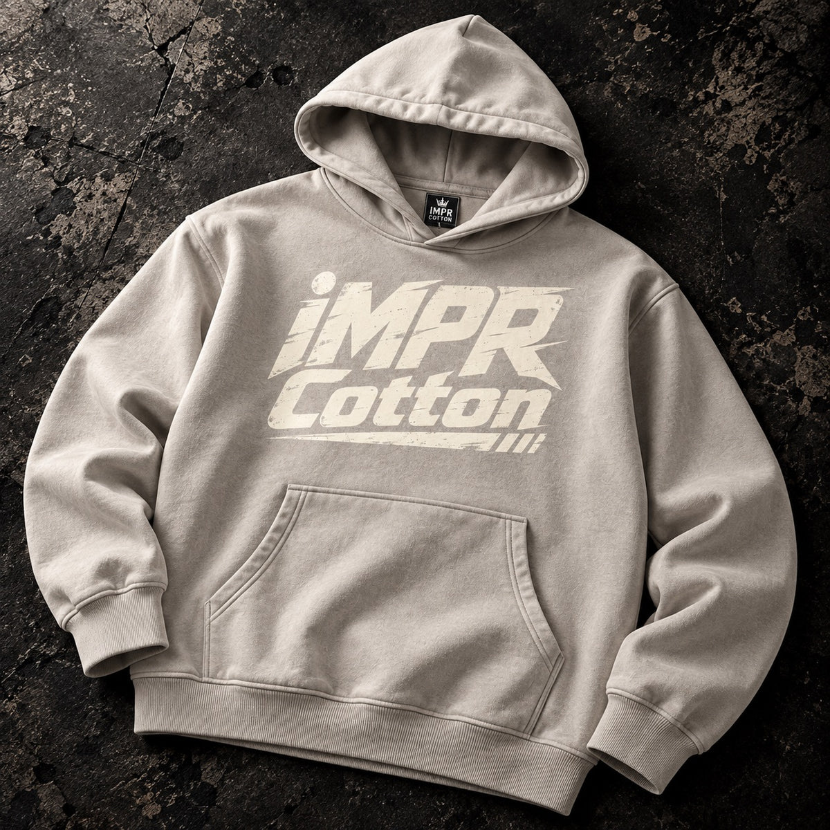 A light beige pullover hoodie with a distressed white graphic reading 'iMPR Cotton' across the chest, laid flat on a dark textured background.