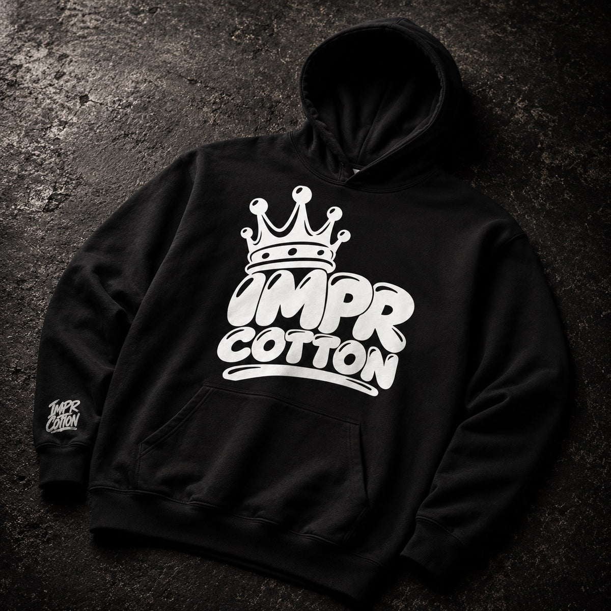 A black pullover hoodie laid flat on a dark textured surface, featuring a large white graphic on the chest with a crown above the words “OMP R COTTON.” A smaller matching white graphic appears on the left sleeve.