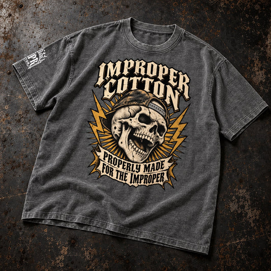 A dark gray acid-wash graphic t-shirt laid flat on a textured dark brown background. The shirt features a large graphic print of a skull wearing a backwards baseball cap, surrounded by yellow lightning bolts and cream-colored text that reads 'IMPROPER COTTON' and 'PROPERLY MADE FOR THE IMPROPER'.