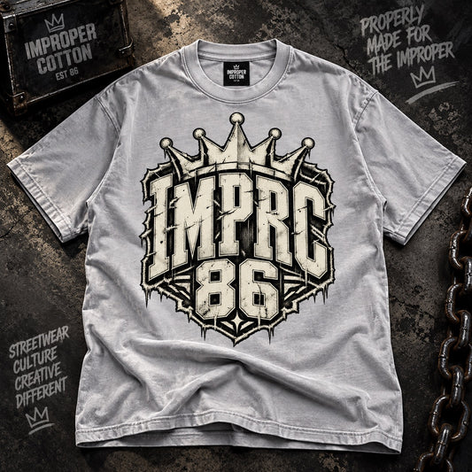 A light gray short-sleeve t-shirt with a large distressed graphic on the front reading 'IMPRC 86' beneath a crown, laid flat on a dark textured background with decorative chain and sign props.