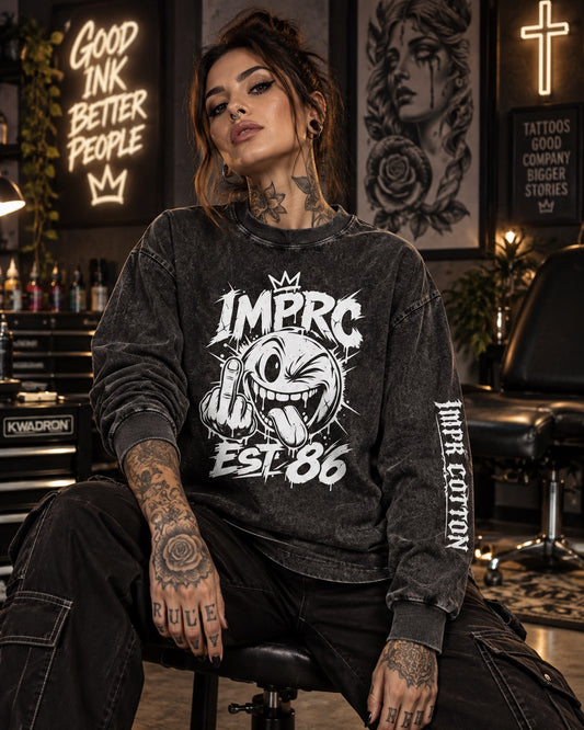 A tattooed woman sits on a stool in a tattoo parlor wearing a dark gray mineral-wash crewneck sweatshirt with a white graphic that reads 'IMPRC EST. 86' and 'IMPR.
