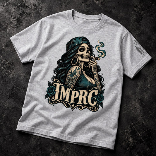 A light heather gray t-shirt laid flat on a dark textured background, featuring a graphic of a Day of the Dead style skeleton woman with a bandana, tattoos, and roses, with the text 'IMPRC' below.