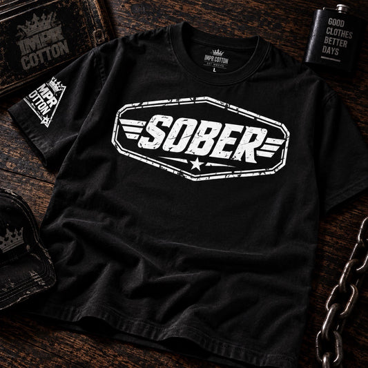 A black short-sleeve graphic T-shirt laid flat on a dark wooden surface, featuring a large distressed white 'SOBER' logo across the chest and a smaller white 'IMP COTTON' logo on the sleeve. Decorative props including a brass bottle, a tin, and a chain surround the shirt.