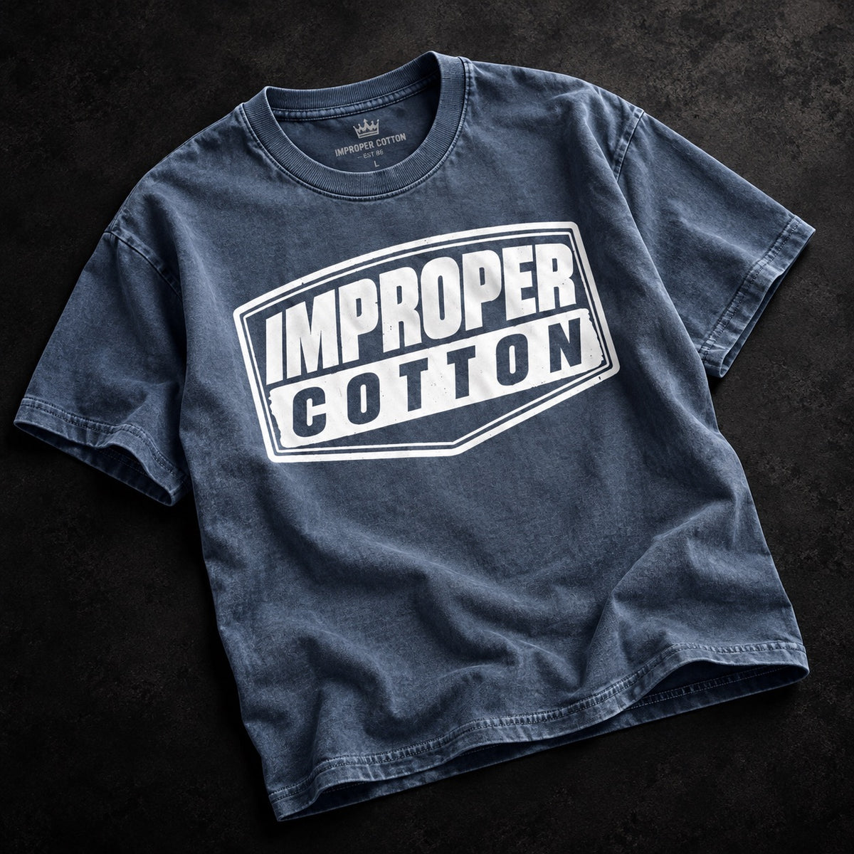 Blue vintage wash IMPROPER COTTON T-shirt displayed at a slight angle on a dark textured background with a bold white signature logo graphic.
