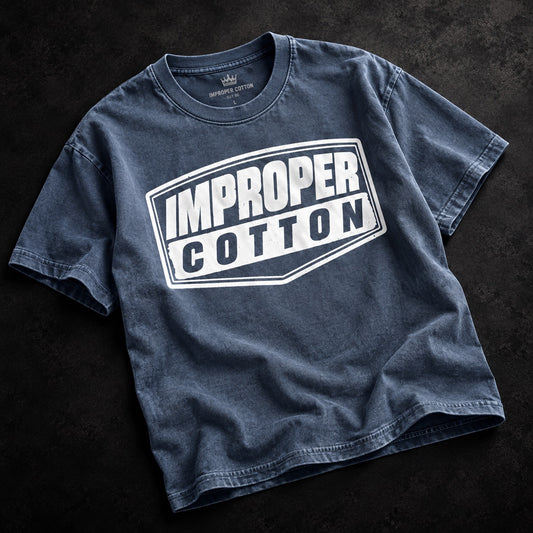 Blue vintage wash IMPROPER COTTON T-shirt displayed at a slight angle on a dark textured background with a bold white signature logo graphic.
