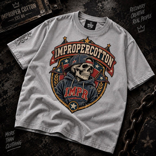 A light gray heather t-shirt with a large graphic print of a skeleton wearing a cap and hoodie, with the text 'IMPROPERCOTTON' and 'IMPR'. The shirt is laid flat on a dark textured background with rustic props.