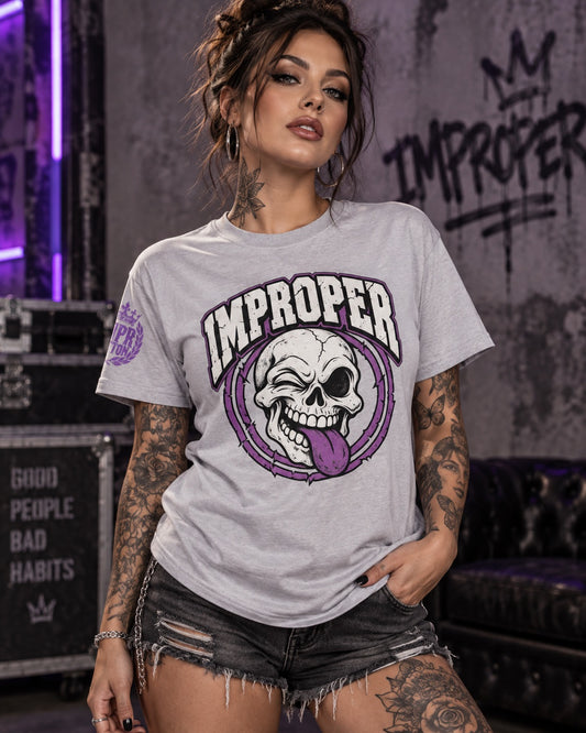 Tattooed female model wearing a light gray IMPROPER graphic T-shirt featuring a winking skull with a purple tongue and distressed purple accents, styled with black ripped shorts in a moody streetwear studio.