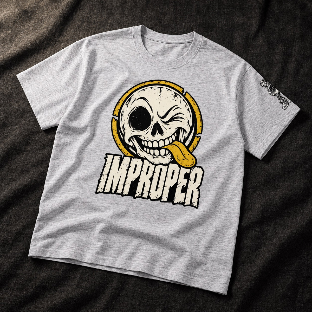 A heather gray t-shirt laid flat on a dark textured fabric background. The shirt features a large graphic print of a winking skull wearing yellow headphones with its tongue sticking out, above the word 'IMPROPER' in a stylized white font with a black outline.
